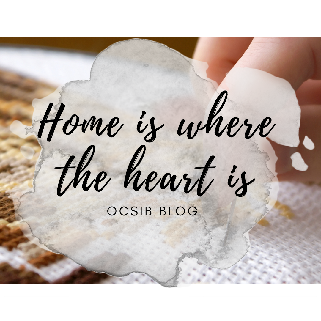 Home is where the heart is OCSIB Blog black text atop water color ink blotch over hand doing cross stitch.