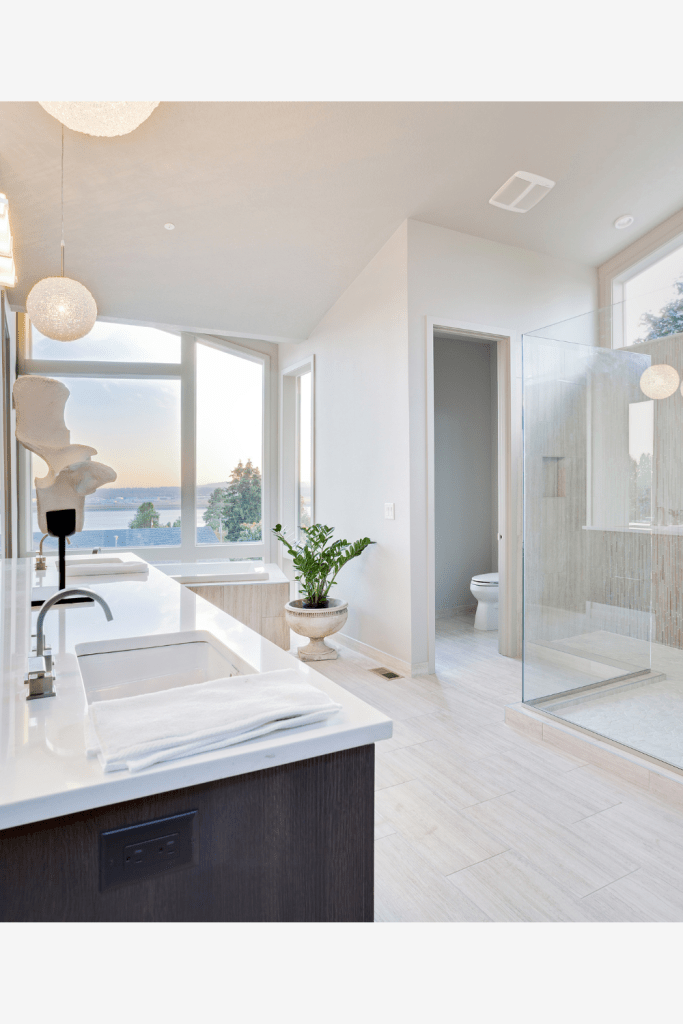 bathroom with white walls, big windows open to view the ocean, espresso cabinets with white stone countertop, beige wood like tile flooring, glass panels and light beige wall tile in shower with crystal ball sphere pendant light and vaulted ceilings