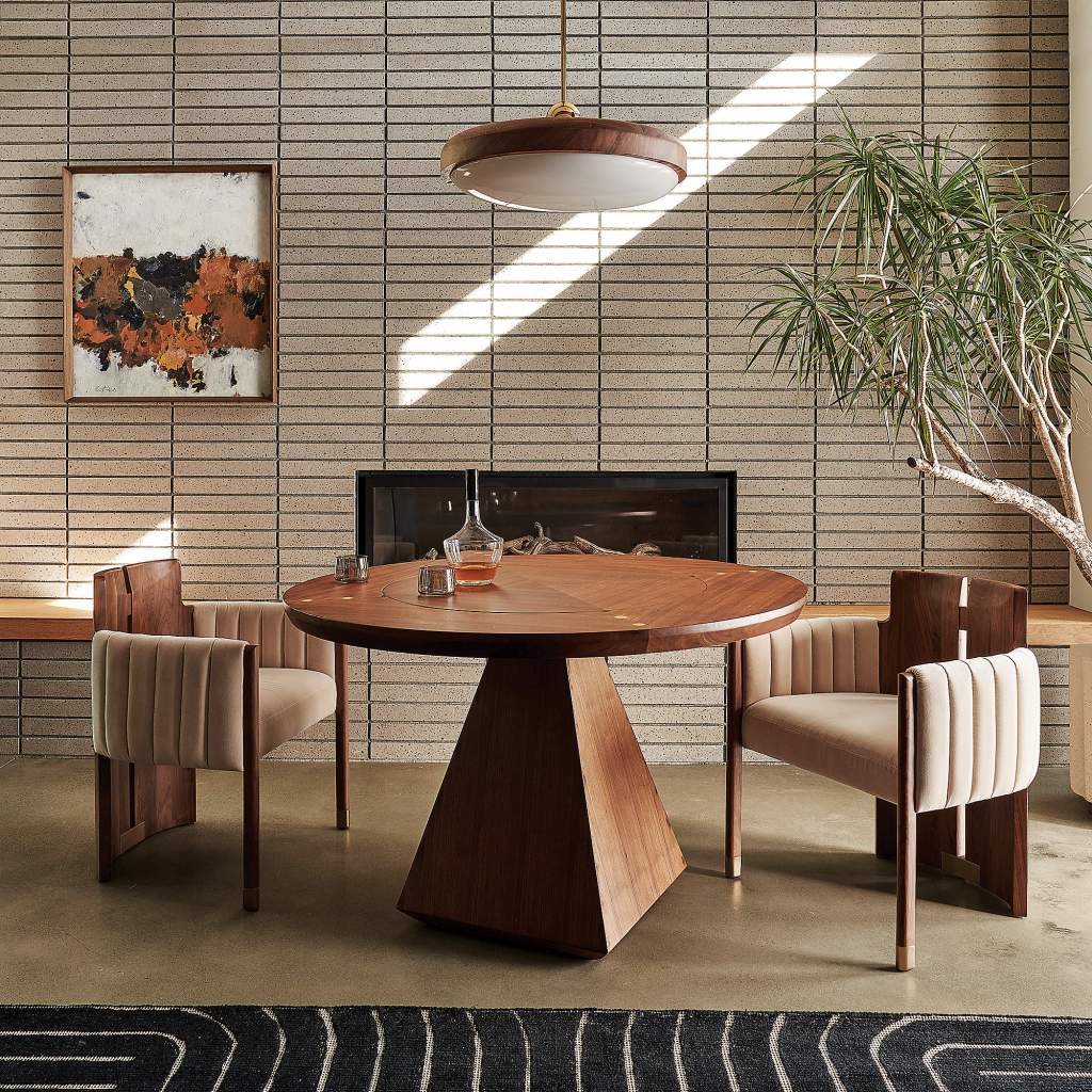 CB2: Capistrano Pendant Walnut wood capped pendant with frosted white glass bowl covering bulbs above a walnut round dining table with beige brick wall behind, fireplace, and two modern walnut wood and beige velvet fabric chairs