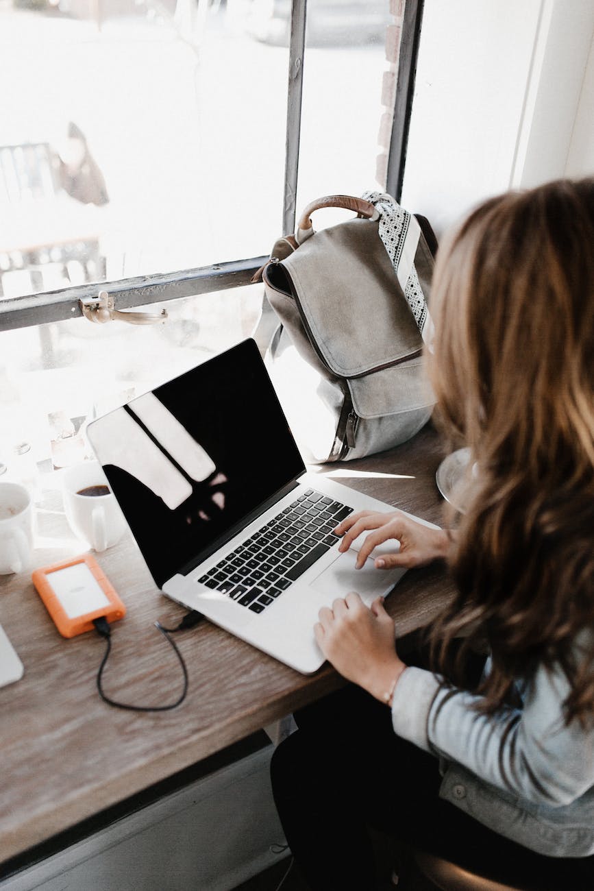woman working on apple laptop at cafe bar with grey purse backpack and a window in front of her. 