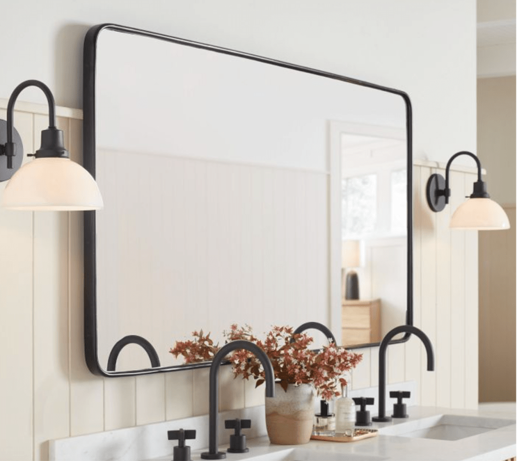 black metal framed rounded corner rectangle mirror long above double sink vanity with black faucets, and black arched wall sconces with bell glass shade