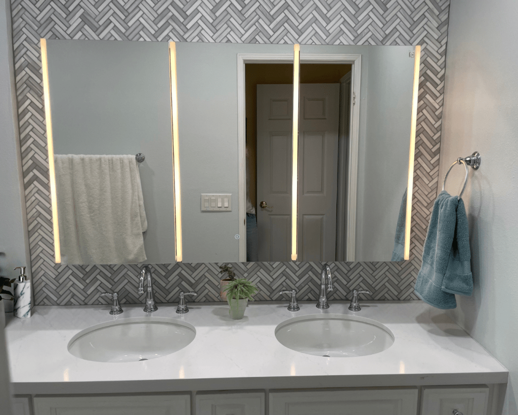 white and grey herringbone mosaic tile on wall 5' wide behind a large three panel lighted mirror above a white quartz countertop two under mount sinks and nickel widespread faucets