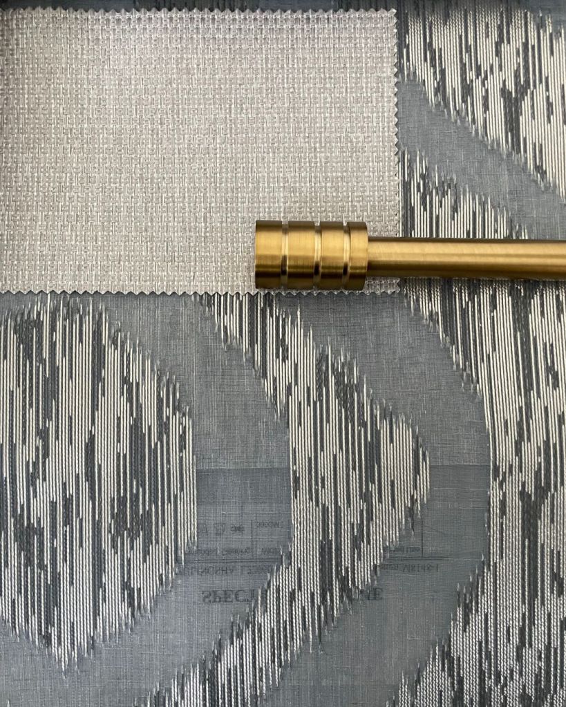 A close-up image of fabric swatches and a curtain rod. The fabric swatches feature a grayish pattern, and the curtain rod is gold-toned.